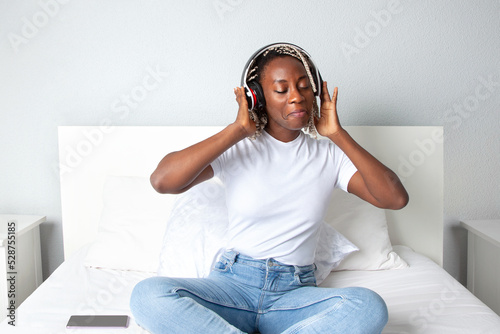 Mujer negra sonriente escuchando música con auriculares en la habitación de su casa. Lifestyle. photo
