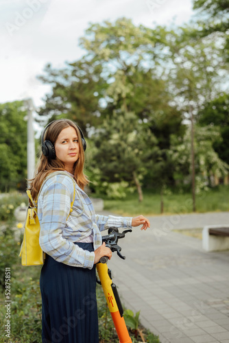 portrait of a young woman with an electric scooter in headphones and with a yellow backpack in the summer on a city street
