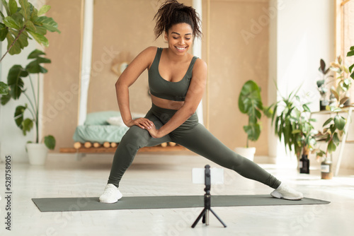 Obraz na plátne Happy fit black lady in sportswear doing side lunge exercise and looking at smar