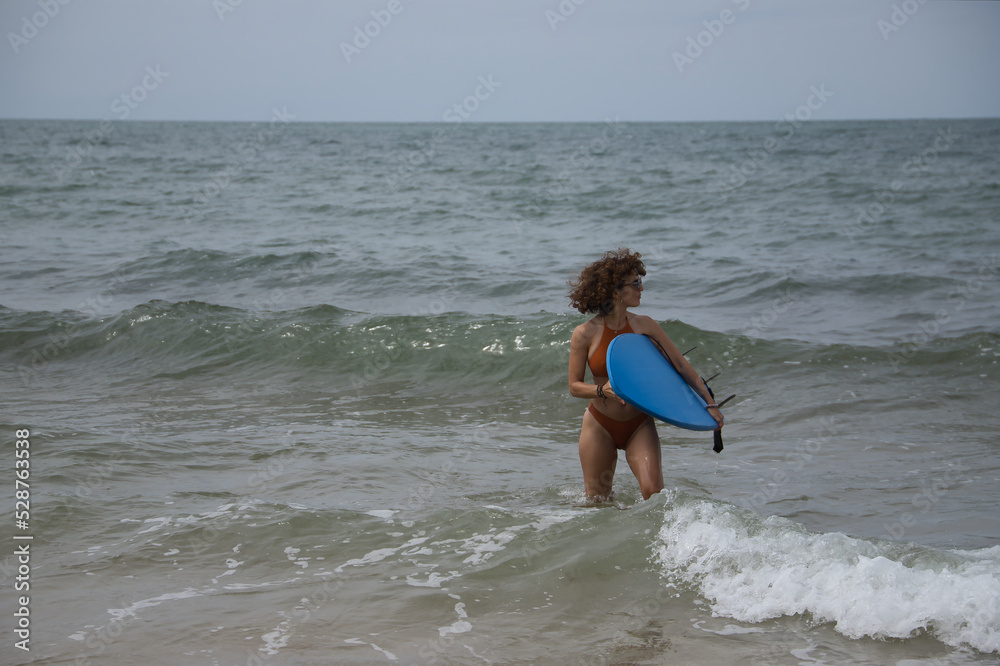 Attractive mature woman with curly hair, sunglasses and bikini, coming out of the water holding a blue surfboard under her arm. Concept sea, sand, sun, beach, vacation, surf, summer. Attractive mature woman with curly hair, sunglasses and bikini, coming out of the water holding a blue surfboard under her arm. Concept sea, sand, sun, beach, vacation, surf, summer.