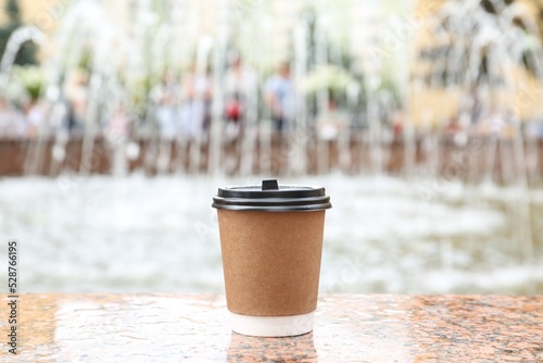 Cardboard cup with plastic lid on stone parapet near fountain outdoors. Coffee to go