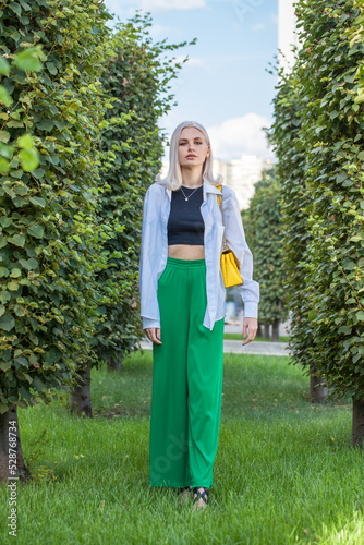 Portrait of a young beautiful blonde girl in green trousers posing in a summer garden