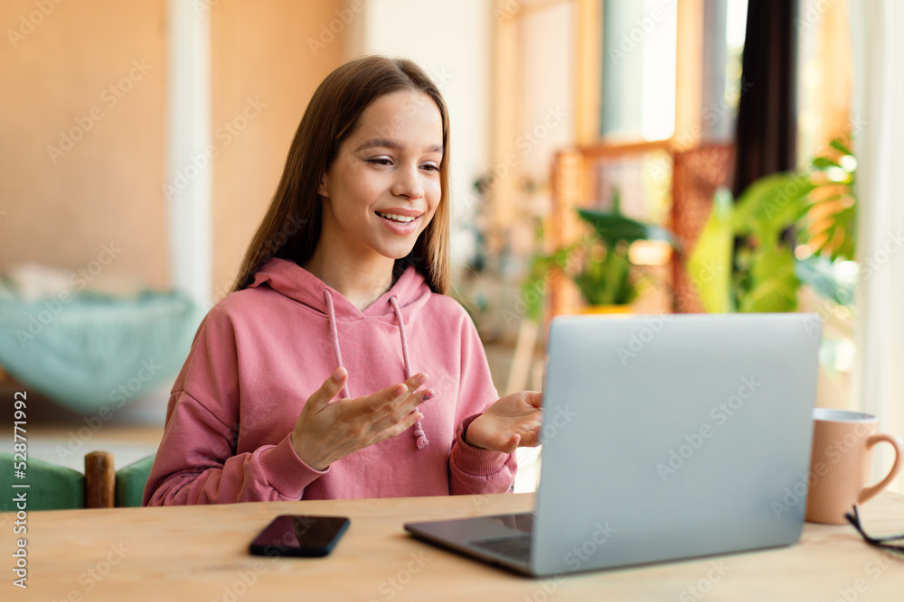 Positive teen girl video calling on laptop and gesturing at webcamera, sitting at desk at home Positive teen girl video calling on laptop and gesturing at webcamera, sitting at desk at home