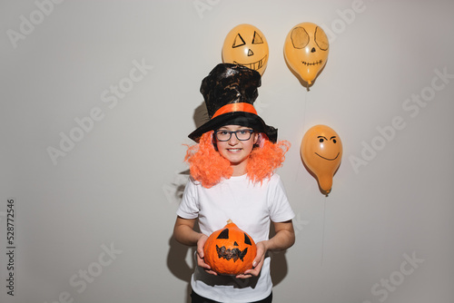 Happy Halloween. Child in orange wig and black hat is holding pumpkin with painted face. The girl is smiling and wearing glasses. Autumn, cheerful mood. All Saints' Day. Soft Selective focus