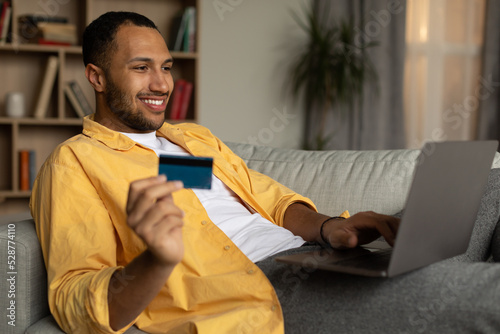Remote shopping. Cheerful black man in headphones buying things on web, using laptop and credit card in living room