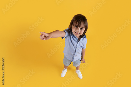 Top view portrait of cute little boy pointing aside at free space and smiling, yellow studio background
