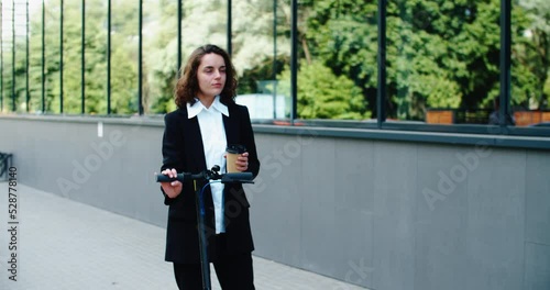 Young smiling businesswoman in a business suit while riding an electric scooter drinking coffee on the background of the shopping center. High quality 4k footage
