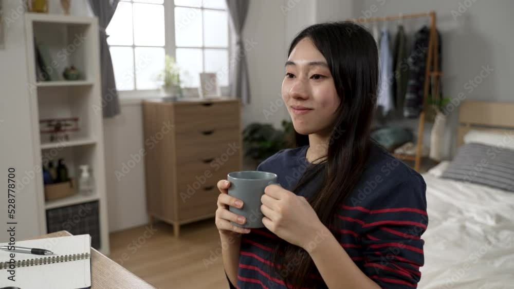 attractive Asian college girl learning from home is smiling into distance while enjoying tea and the tranquil morning at desk in her modern bedroom with daylight attractive Asian college girl learning from home is smiling into distance while enjoying tea and the tranquil morning at desk in her modern bedroom with daylight