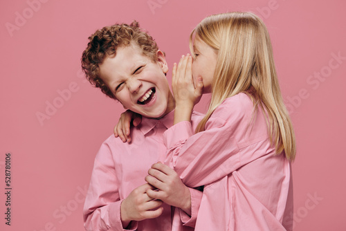 cute, beautiful, happy children are standing in pink clothes on a pink background and the girl tells the boy secrets in his ear. Topics of friendship and relationships