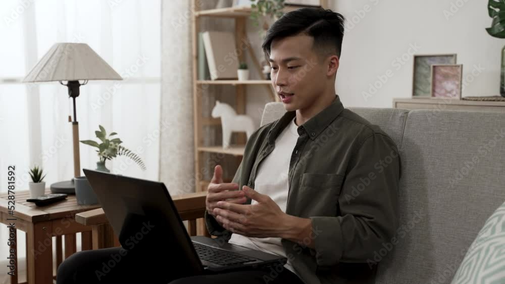 asian male creative worker is waving and explaining to his client with hand gestures while having an online meeting on the computer in the living room at home. asian male creative worker is waving and explaining to his client with hand gestures while having an online meeting on the computer in the living room at home.
