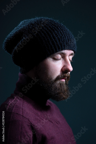 bearded guy with closed eyes in a knitted black hat and burgundy sweater. On a gray background