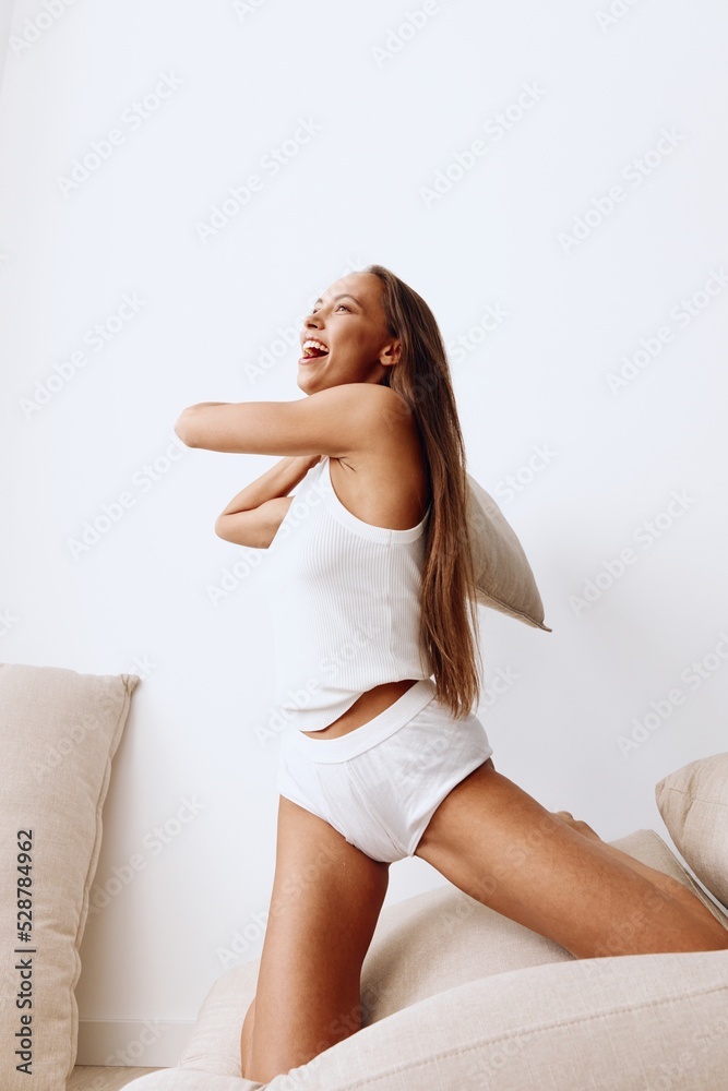 Beautiful woman in homemade white lingerie plays with couch cushions throws up with a smile and has fun. Lifestyle in a happy home and fun time Beautiful woman in homemade white lingerie plays with couch cushions throws up with a smile and has fun. Lifestyle in a happy home and fun time