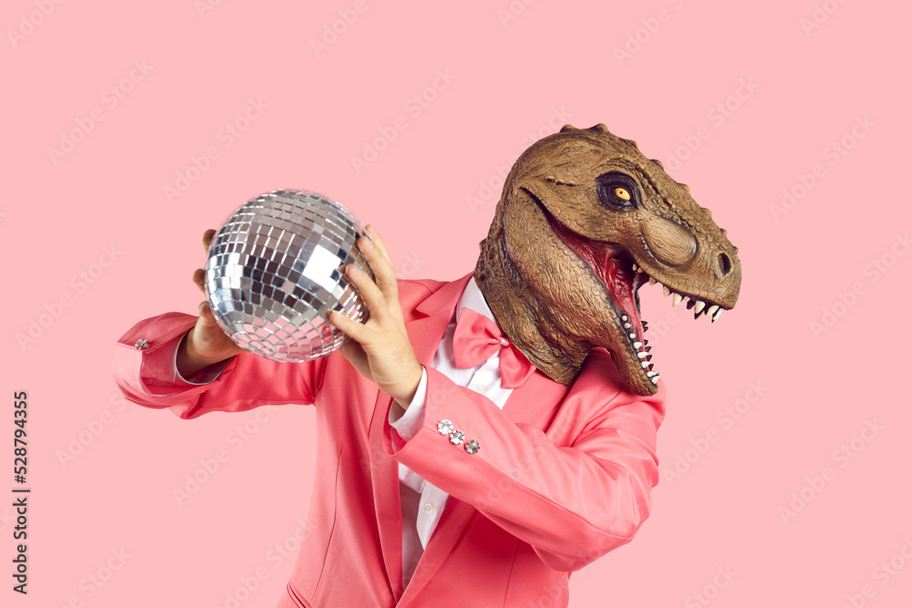 Man in rubber dinosaur head mask isolated on pink background wear costume  dance holding disco ball. Smiling male entertainer in lizard headgear have  fun on party or celebration. Carnival concept. Photos