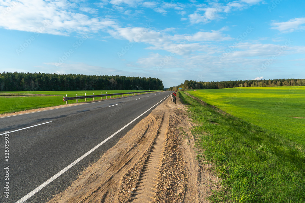 An empty roadway outside the city passes by a rural field and a blooming meadow. Travel by car away from the city and the hustle and bustle. Paved road on a sunny day without cars. An empty roadway outside the city passes by a rural field and a blooming meadow. Travel by car away from the city and the hustle and bustle. Paved road on a sunny day without cars.