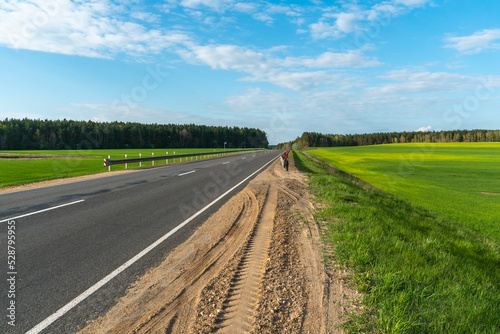 An empty roadway outside the city passes by a rural field and a blooming meadow. Travel by car away from the city and the hustle and bustle. Paved road on a sunny day without cars.