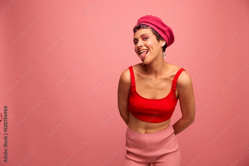 Young athletic woman with a short haircut and purple hair in a red top and a pink hat with an athletic figure smiles and grimaces looking at the camera on a pink background Young athletic woman with a short haircut and purple hair in a red top and a pink hat with an athletic figure smiles and grimaces looking at the camera on a pink background