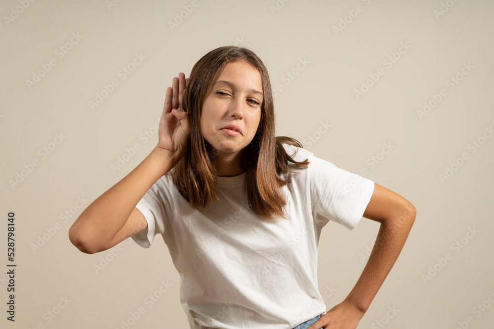 Excited teen girl holding hand next to ear listening to something over beige background. Confidential girl listens to news or gossip copy spaced on isolated background. Advertising shopping concept. Excited teen girl holding hand next to ear listening to something over beige background. Confidential girl listens to news or gossip copy spaced on isolated background. Advertising shopping concept.
