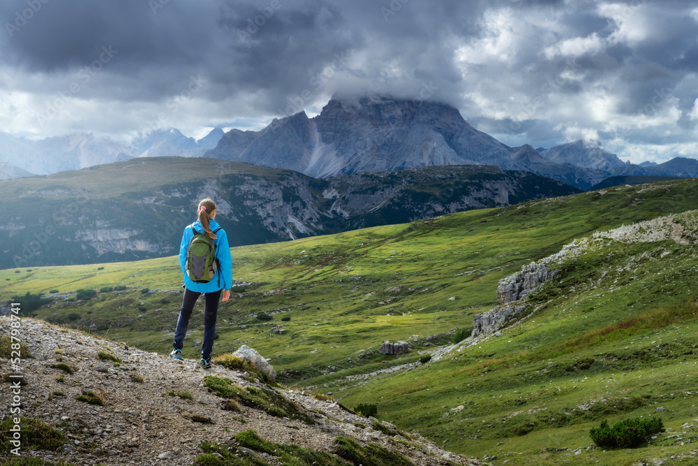 Girl with backpack on the hill looking at mountains at sunset in summer. Dramatic landscape with young woman, high rocks, trail, green grass, cloudy sky in Dolomites, Italy. Adventure and hiking