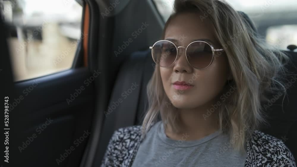 Attractive asian young woman travels by car, sitting on back seat of taxi, looking out window. Beautiful girl wearing sunglasses uses transfer service in journey Attractive asian young woman travels by car, sitting on back seat of taxi, looking out window. Beautiful girl wearing sunglasses uses transfer service in journey