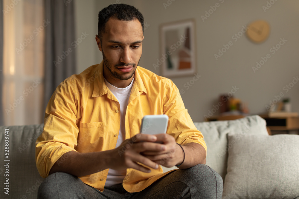 Serious millennial black man using cellphone, messaging online, sitting on sofa at home, copy space Serious millennial black man using cellphone, messaging online, sitting on sofa at home, copy space
