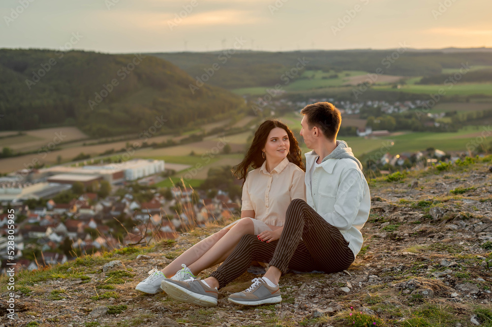 A couple in love, a guy and a girl are sitting on a mountain at sunset kissing and hugging