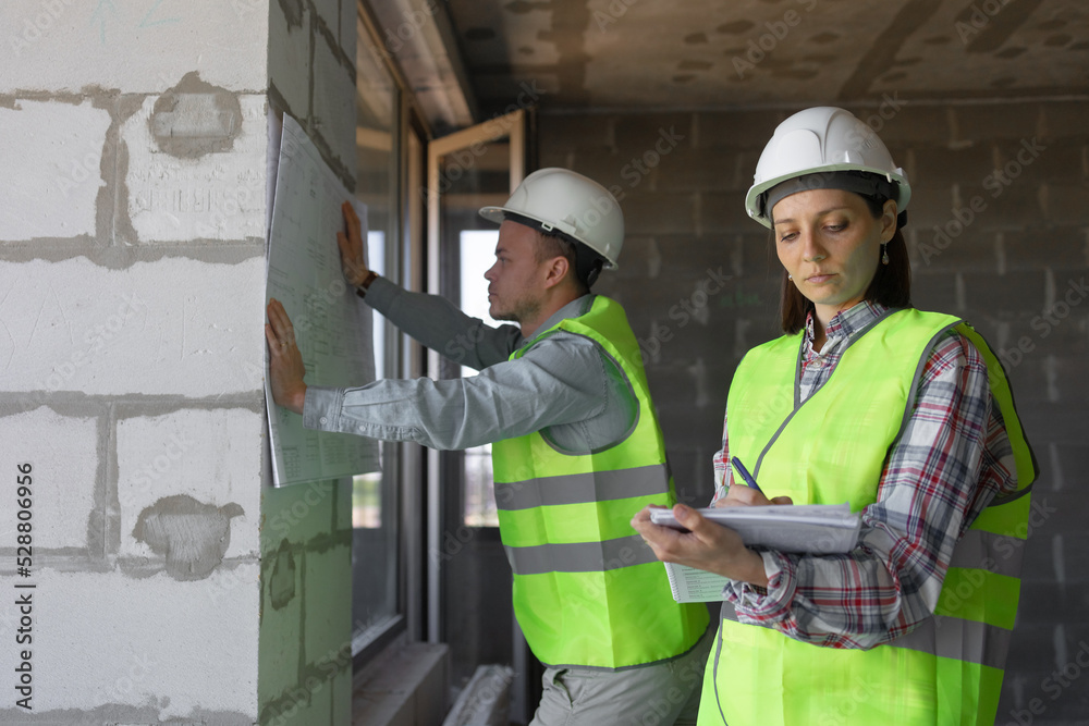 two engineers, a man and a woman in white helmets and protective vests, are standing in the room and holding a plan of the construction site and fire safety in their hands. interior designers