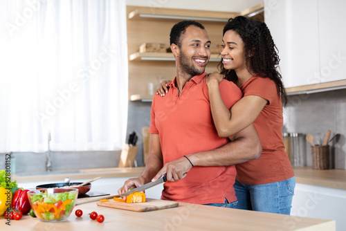 Happy young black couple in same t-shirts prepare homemade meal, wife hugs husband, man cuts vegetables
