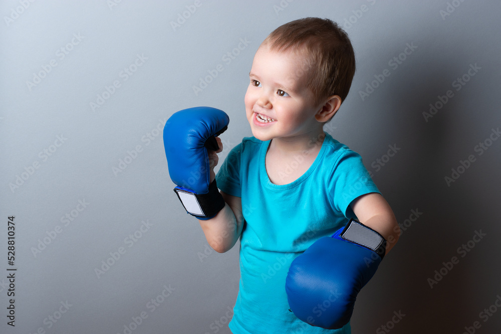 a little boy in boxing gloves and a blue T-shirt on a gray background.expression of emotions.copy space. a little boy in boxing gloves and a blue T-shirt on a gray background.expression of emotions.copy space.