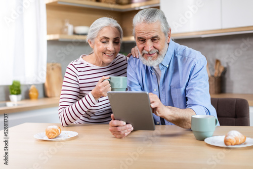 Happy Senior Spouses Using Digital Tablet Together While Having Breakfast In Kitchen
