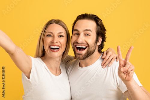 Glad happy millennial caucasian husband and wife in white t-shirts have fun, make peace sign and selfie
