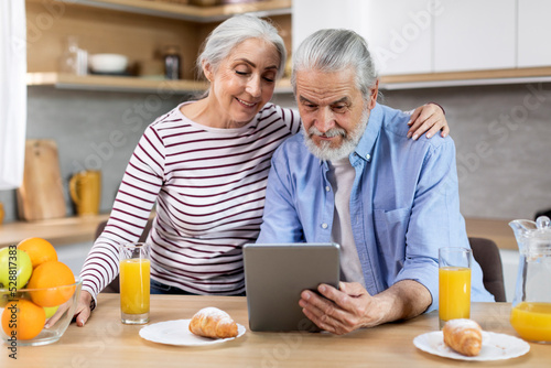 Happy Senior Couple Browsing Internet On Digital Tablet During Breakfast In Kitchen