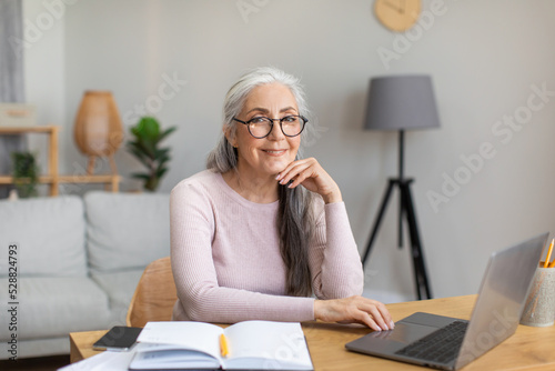 Smiling glad caucasian old gray-haired lady in glasses typing on laptop, chatting in living room interior