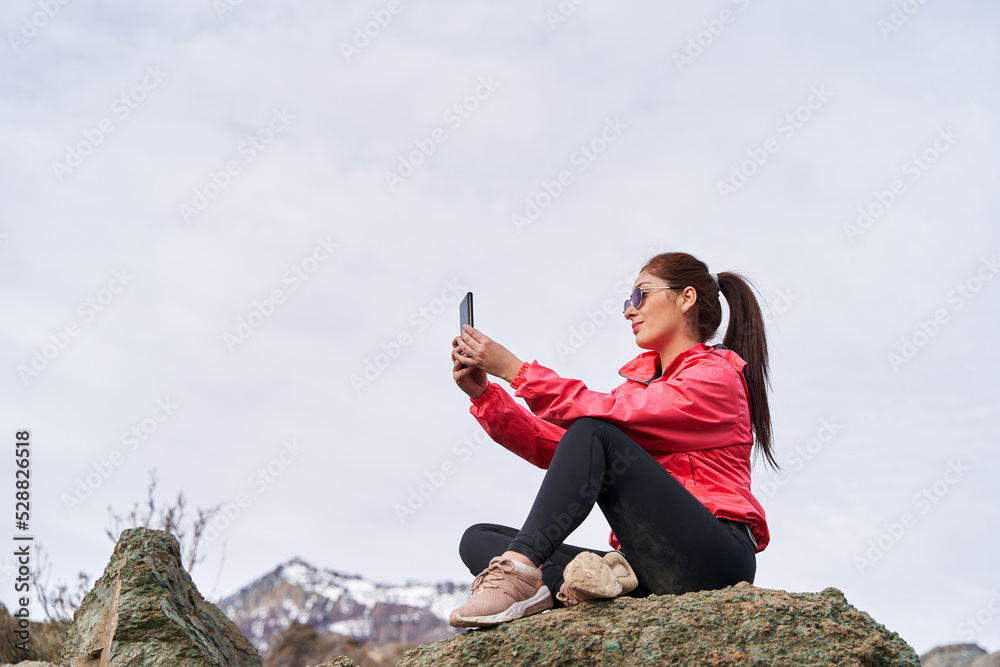young woman with red hair, taking a selfie, wearing a red jacket, sitting on a rock in the middle of the Andes mountain range in Chile young woman with red hair, taking a selfie, wearing a red jacket, sitting on a rock in the middle of the Andes mountain range in Chile