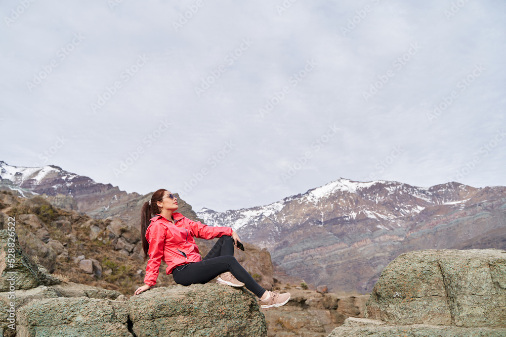 young red haired woman with her hair tied up, wearing a red jacket, sitting on a rock in the middle of the Andes Mountains of Chile