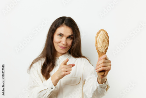 beautiful cute pleasant woman with long black hair stands holding a wooden comb in her hands and points her finger at her smiling cutely. Horizontal photo on a light background