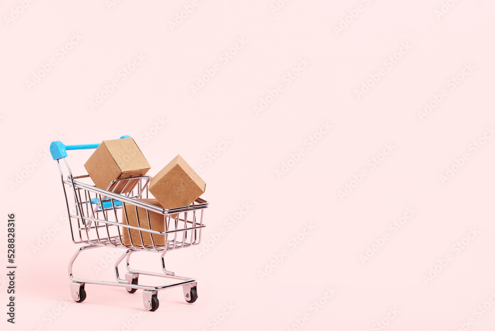 Shopping cart with cardboard boxes on a pastel pink background. Bright minimalist design with copy space. Concepts: market sales, seasonal discounts, logistics, packaging.