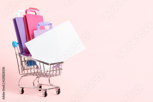 Shopping cart loaded with paper bags and a blank card on a pastel pink background. Minimalist design with copy space. Concepts: market deals, seasonal sales and discounts, black friday.