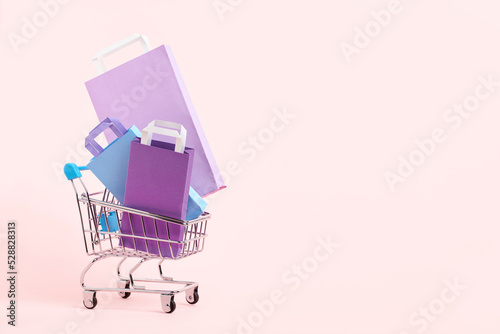 Shopping cart with purple, lilac and blue paper bags on a pastel pink background. Minimalist design with copy space. Concepts: market deals, seasonal discounts and rebates, black friday sales.