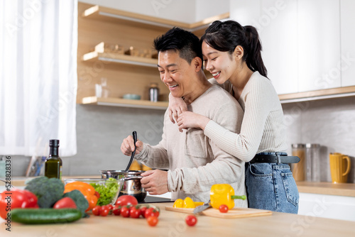 Happy young asian lady hugging her spouse cooking dinner