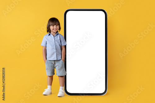 Cute happy little boy smiling to camera, posing near big blank smartphone with hands in pockets, empty space