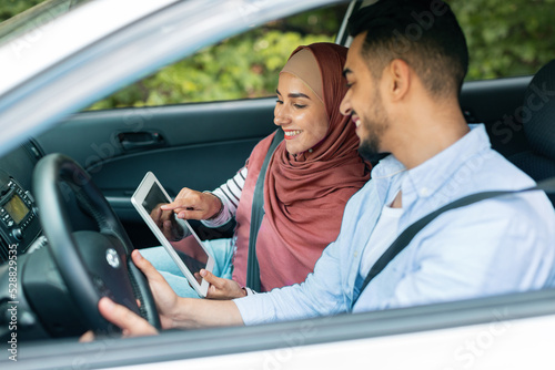 Cheerful millennial muslim female in hijab shows way on tablet with empty screen to husband at steering wheel in car