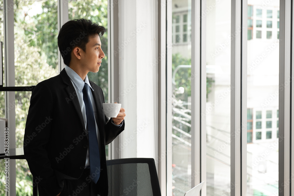 Thoughtful young businessman drinking coffee, looking through window, having break, deep in thoughts, enjoying view, waiting for meeting to start, making business decision.