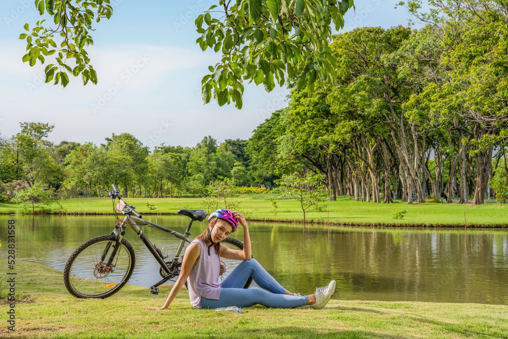 asian female biker sitting beside bicycle by lake in park relaxing after cycling for exercise during summer
