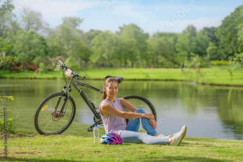 asian female biker sitting beside bicycle by lake in park relaxing after cycling for exercise during summer