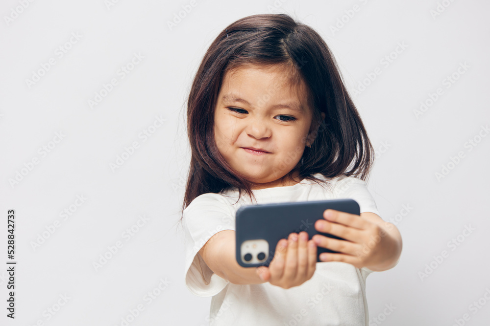 cute, funny, little girl in a white t-shirt stands on a light background with a smartphone in her hand and takes a selfie. Horizontal studio photography with blank space for advertising mockup insert cute, funny, little girl in a white t-shirt stands on a light background with a smartphone in her hand and takes a selfie. Horizontal studio photography with blank space for advertising mockup insert