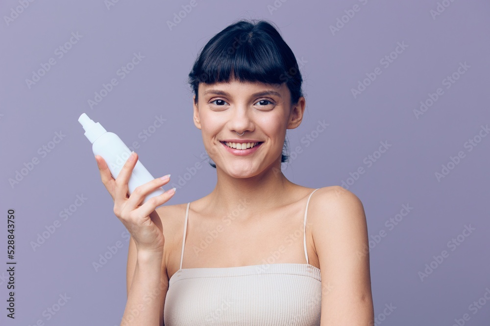 Horizontal studio shot.A beautiful joyful woman in a white T-shirt with clean beautiful well-groomed skin, black wavy short hair gathered in a ponytail stands on a lilac background with a bottle Horizontal studio shot.A beautiful joyful woman in a white T-shirt with clean beautiful well-groomed skin, black wavy short hair gathered in a ponytail stands on a lilac background with a bottle