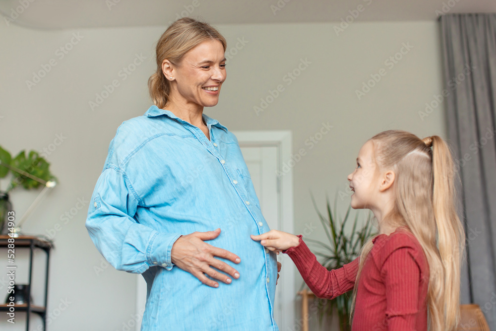Fototapeta premium Happy little girl touching pregnant mother's belly, spending time at home together, looking at each other and smiling