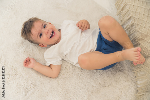 Amused caucasian preschooler boy in white t-shirt and dark-blue short lying on his back on the bed and laughing.Happy childhood concept. High quality photo