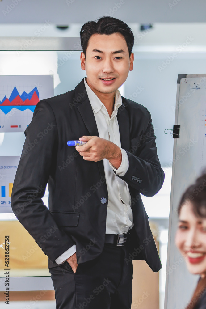Asian handsome professional successful businessman presenter standing holding pen pointing presenting company graph chart paperwork document on glass board to male female colleagues in meeting room Asian handsome professional successful businessman presenter standing holding pen pointing presenting company graph chart paperwork document on glass board to male female colleagues in meeting room