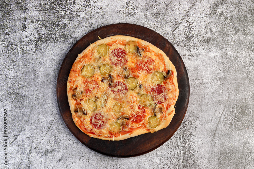 Homemade rustic pizza with pickled cucumbers, cheese and mushrooms on a round wooden cutting board on a dark grey background. Top view, flat lay Homemade rustic pizza with pickled cucumbers, cheese and mushrooms on a round wooden cutting board on a dark grey background. Top view, flat lay